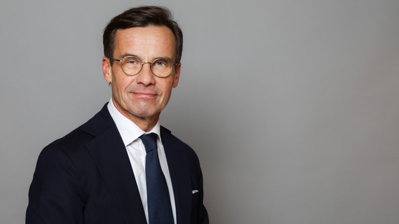 Vit medelålders man, kort brunt bakåtkammat hår i sidbena. Rundade stålbågade glasögon. Mörkblå kavaj, vit skjorta, blå slips.