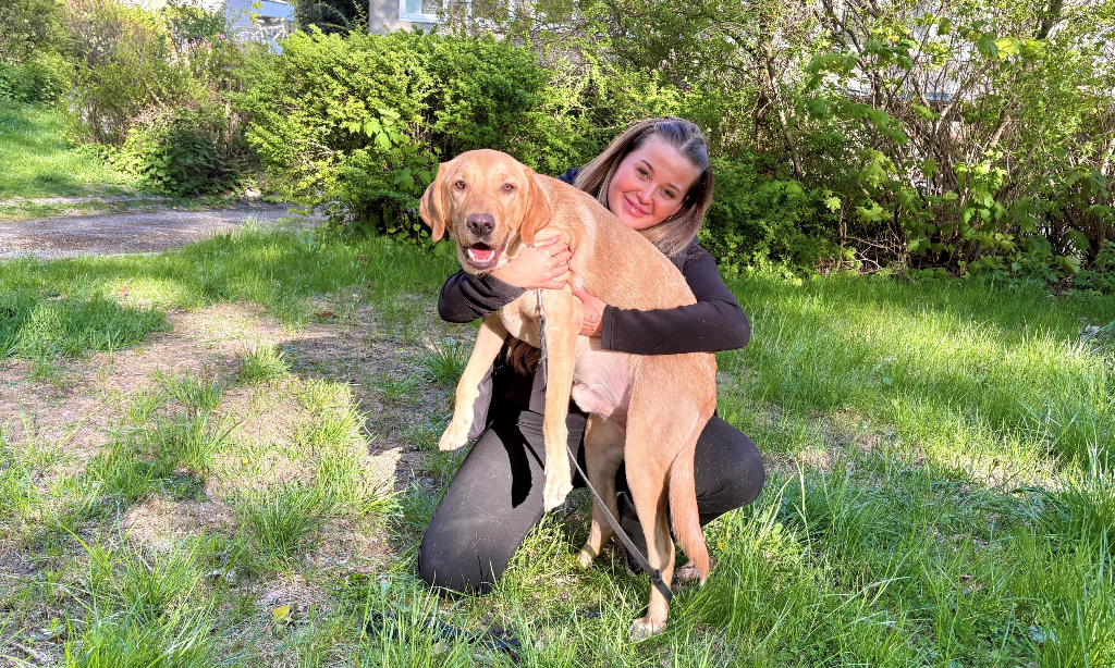 20 årig vit leende tjej med långt rågblont hår och svarta kläder sitter på huk på gräsmatta. Hon har en ljus labrador i knät.
