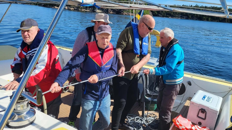 Fem vita män personer står i sittbrunnen på en segelbåt, tre och samarbetar där de drar i en tamp. De bär flytvästar och omges av lugnt vatten, blå himmel och en skogsklädd kust i bakgrunden.