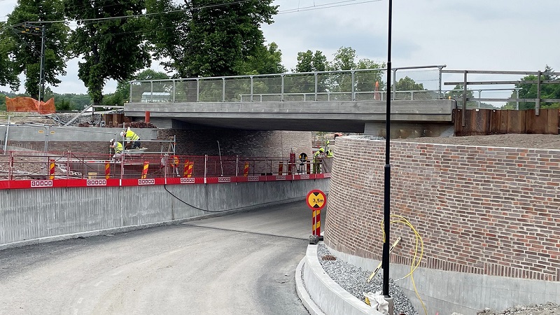 En svängd vägg leder in i en tunnel av ljusbrunt tegel i varierande nyanser. Ovanför går en järnvägsbro.