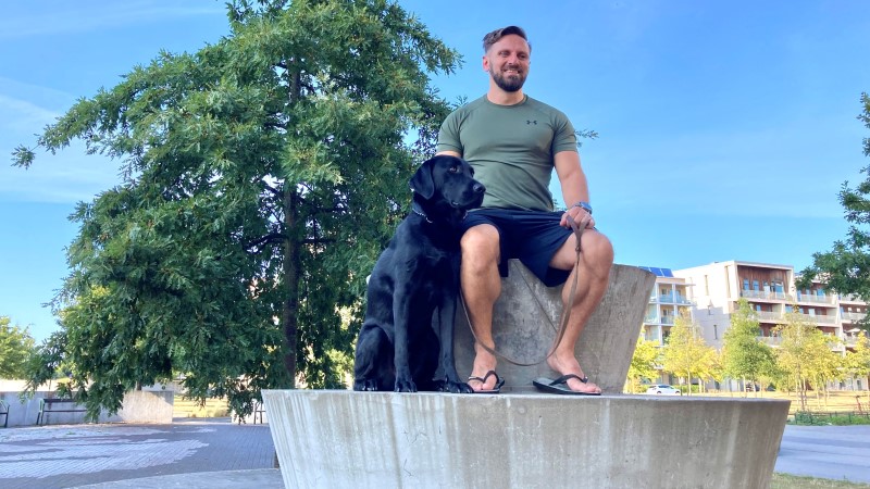 En medelålders vit man med kort brunt hår, kakigrön t-shirt, mörkblåa shorts. Sitter på en rund trappformad cementskulptur. Bredvid sig hans svarta labrador-ledarhund. En solig dag, blå himmel. Stort träd bakom.
