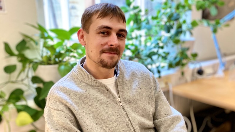 Vit 30-årig man sitter i en stol. Kort rakt brunt hår, mustache och bockskägg. Ljusgrå stickad kofta. Gröna krukväxter i bakgrunden..