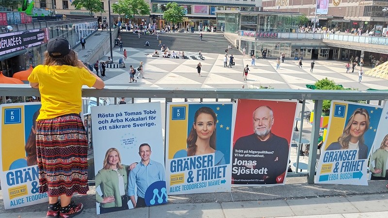 En vy över Sergels Torg, i förgrunden syns olika partiers valaffischer. En kvinna i gul t-shirt och flerfärgad kjol står med ryggen till och ser ut över torget.