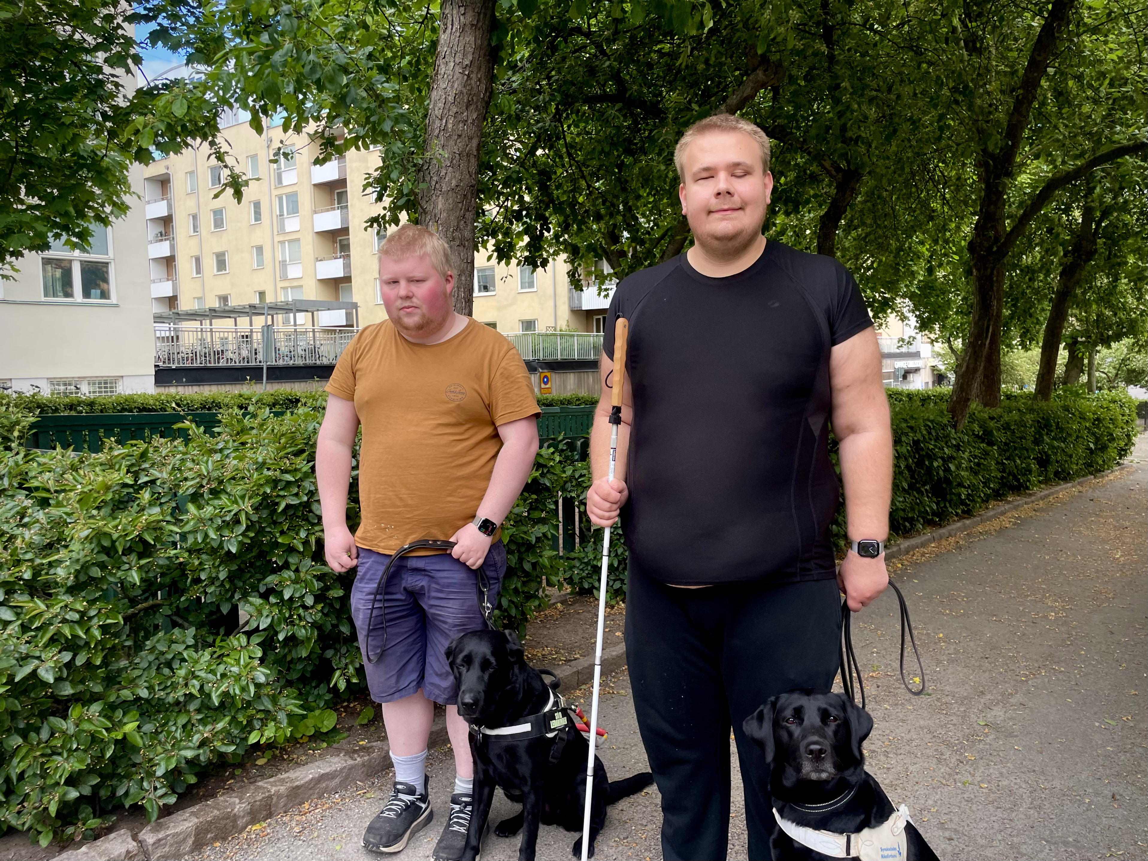 Två vita unga män står tillsammans med sina svarta ledarhundar. Till vänster Oliwer: som har en orange t-shirt och blå shorts. Kevin till höger: har en svart t-shirt och svarta långbyxor. Båda har ljust kortklippt hår. De står i en grönskande trädallé. Kevin håller i sin vita käpp.