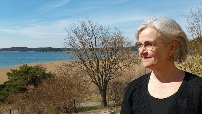 En vit kvinna i gråhårig page och glasögon ser ut över en havsvik.