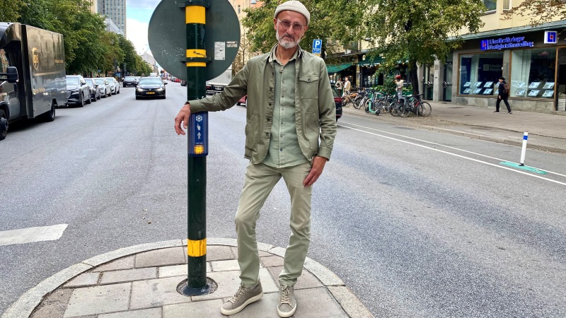 Medelålders vit solbränd man står vid refug i mitten av gata. Han har kakigrön jacka,  ljusare byxor. Beige liten mössa. Vitt skägg och glasögon.