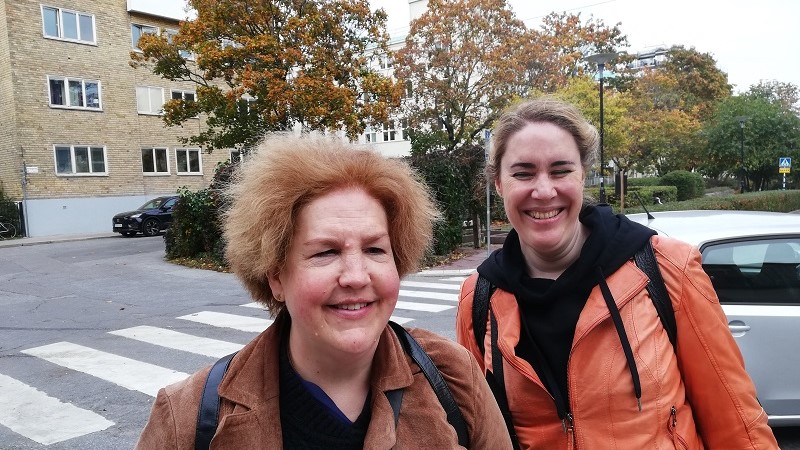 Vänster: vit medelålders kvinna, rödbrunt burrigt hår, har brun mockajacka. Till hö: vit yngre kvinna, har uppsatt mörkblont hår, en orange läderjacka, svart huvtröja under. De står framför ett övergångsställe vid en park.