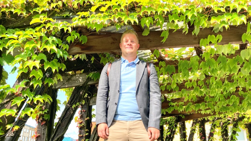 Ung vit man i grå kavaj, ljusblå tröja står under grönskande spaljetak.