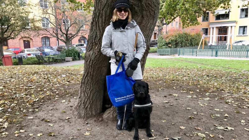 Kvinna står vid ett stort träd i park, svart ledarhund bredvid