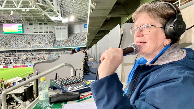 En vit medelålderskvinna, i mörkblå jacka, har mikrofon och hörlurar, sitter på en fotbollsläktare och syntolkar. Grön fotbollsplan med kvinnliga spelare i bakgrunden.