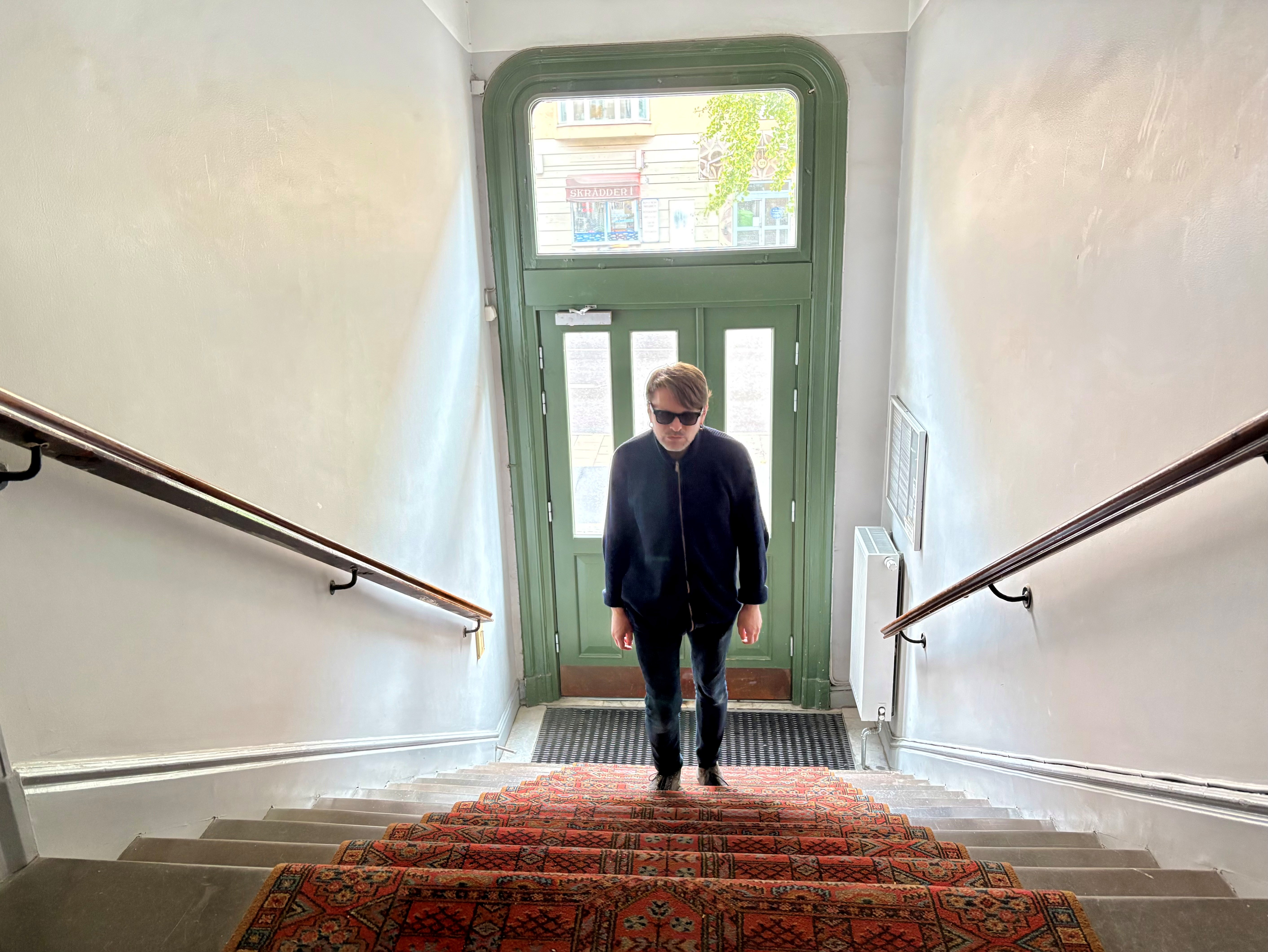En slank 51-årig vit man med kort brunt hår och mörka glasögon, går upp för en trappa i en äldre trappuppgång. Porten är grön, med utsikt mot gata. Han har svart jacka och byxa