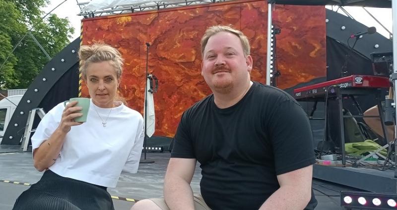 På en scenkant utomhus sitter en blond kvinna med en kaffekopp i handen och en mörkblond man med rödlätt mustasch och litet skägg.