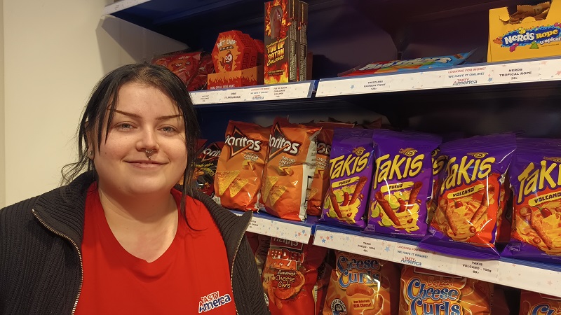 En ung ljushyad kvinna (Alethea) med mörkt uppsatt hår framför påsar med olika sorters snacks med stark kryddning.