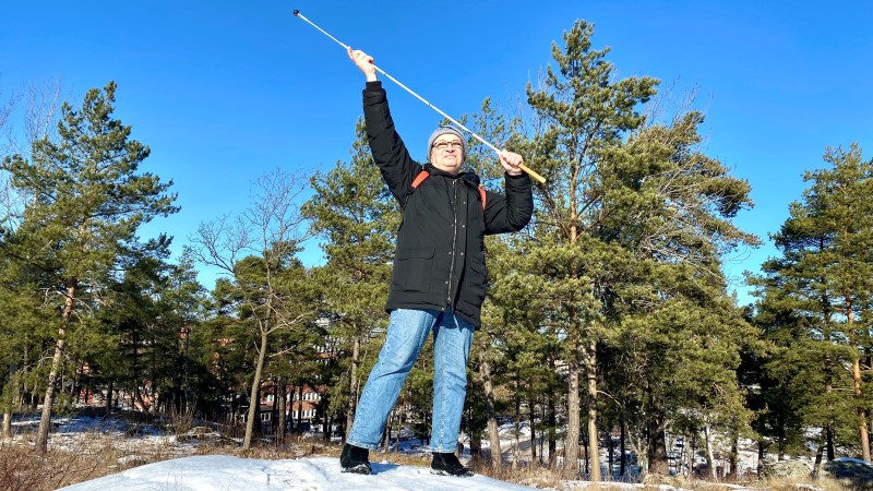 Medelålders vit man med mörkt hår och tjocka glasögon står på en snökulle. Han har mörkblå jacka och blåjeans. Håller upp sin vita käpp med händerna över huvudet.