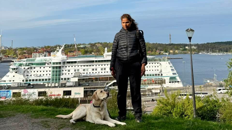 En 40-årg kvinna med långt mörkbrunt hår, står på ett berg med utsikt över vatten, stor vit färja i bakgrunden. Bredvid sig har hon ljus labrador, ledarhund.