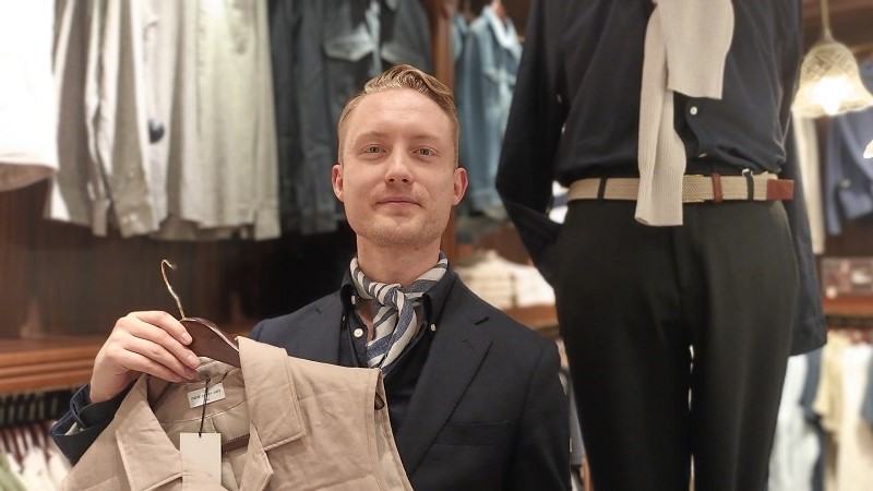 En ung man med blond vågig lugg snett bakåtkammad, iklädd mörkblå kavaj och mörk skjorta och en blåvit halsduk knuten vid kragen. Han håller upp en beige väst.