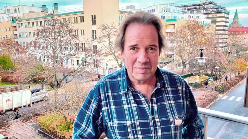 Medelålders vit man, brunt kort hår, bruna ögon, mörkt blåvitrutig skjorta. Står på balkong med gatuvy.
