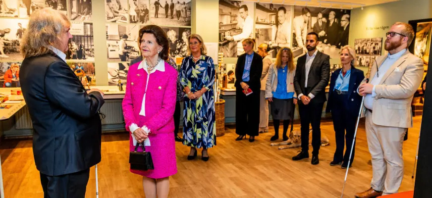 Drottningen i ceriserosa dräkt står med en grupp människor i ett av museirummen. På väggarna stora svartvita foton av personer som arbetar med hantverk för blinda.