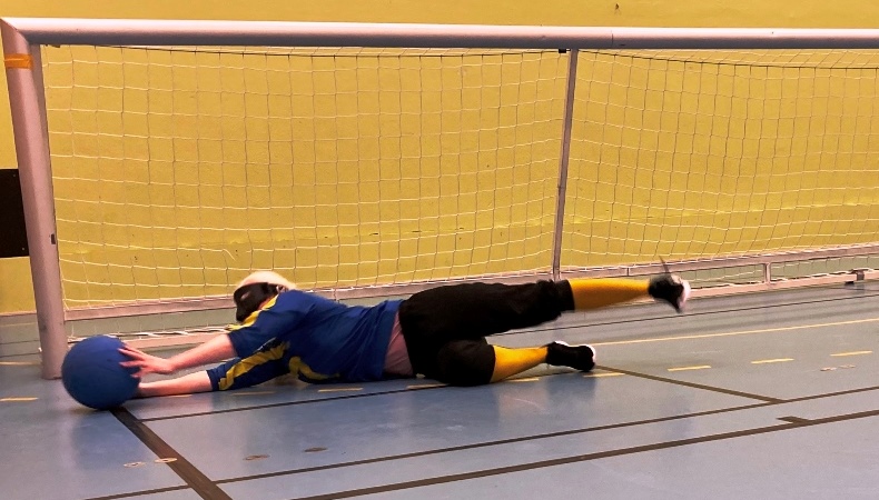 Blond liggande kvinna tar emot en blå boll framför ett Goalballmål