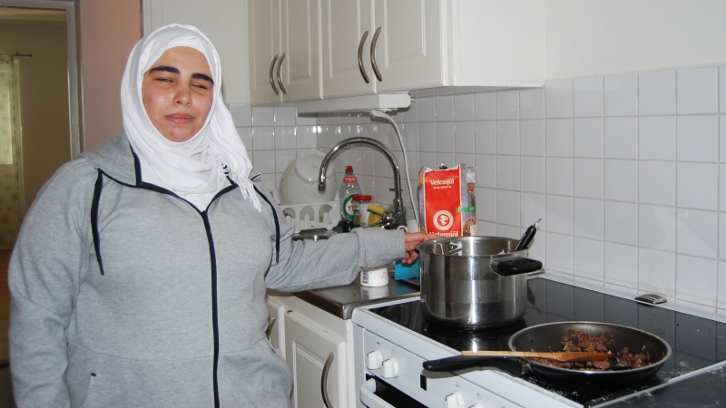 En ung kvinna i vit huvudduk och grå träningsjacka, står vid spis med kastrull och stekpanna på. Foto: Sophie Heine