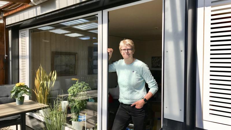 Blond kortklippt kvinna med glasögon lutar sig mot en dörren till en altan.