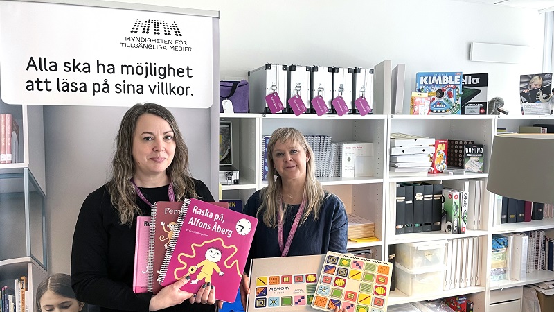 Ida Bengmark med brunt vågigt hår och Catinka Grunditz blond med rakt hår och lugg visar upp material för barn med synnedsättning. En Alfons Åberg-bok och ett memoryspel. Bakom Ida står texten ”Alla ska ha möjlighet att läsa på sina villkor”.