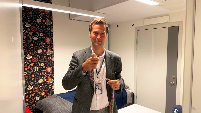 Medelålders vit man kort brunt hår, grå kavaj står i ett läkarundersökningsrum.