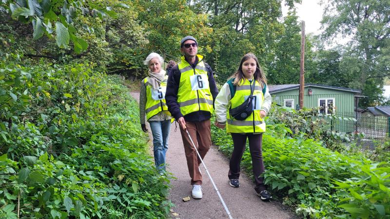 På en stig omgiven av grönska vandrar tre personer med neongrön väst och startnummer 5, 6 och 7. Personen i mitten håller en vit käpp.