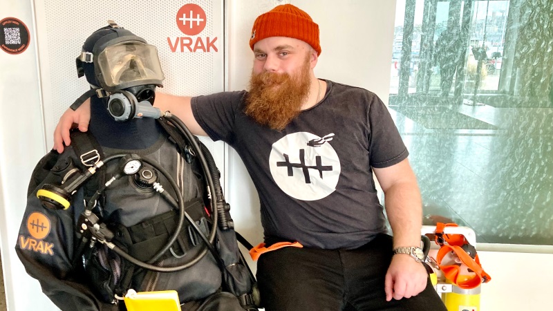 Till höger en vit lite satt man med långt rött helskägg, som har armen om en modell av en dykare i full utrustning. De sitter på en bänk.