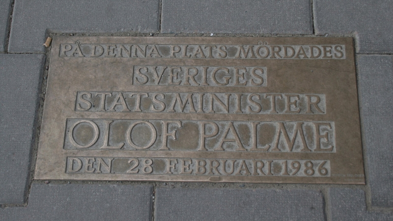 En minnesplakett i brons mott grå gatstenar. Längt upp till vänster i skarven mellan plaketten och gatstenen ligger en cigarettfimp Texten på plaketten lyder ”PÅ DENNA PLATS MÖRDADES SVERIGES STATSMINISTER OLOF PALME DEN 28 FEBRUARI 1986”