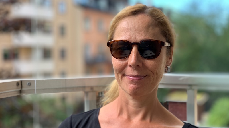 Leende kvinna med solglasögon och rödblont hår samlat i tofs i nacken sitter på en balkong med bostadshus och grönska i oskarp bakgrund. 