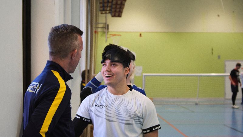 En ung vit man i en vit träningströja pratar med en medelålders vit man i träningsoverallsjacka. Den unge mannen ler och har en svart ögonmask uppdragen i pannan. Platsen är en gymnastikhall och i bakgrunden syns ett goalball-mål.