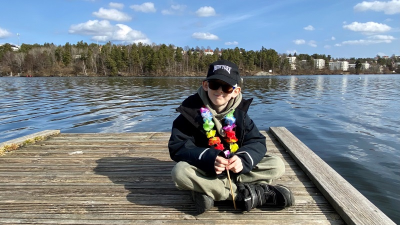 En pojke med blå keps och blå jacka sitter på en solig badbrygga med skogslandskap på andra sidan vattnet.