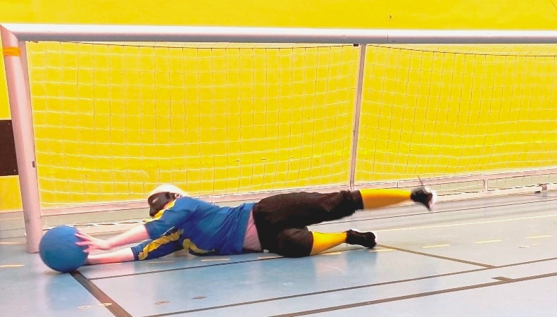 Blond kvinna slänger sig framför ett goalballmål, fångar blå boll