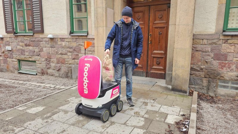 En man står på en gata och lägger ner en kasse med mat i en Foodora-robot, som är en självkörande robot-låda. Mannen har en blå jacka och Foodora-roboten är vit/rosa-röd.  