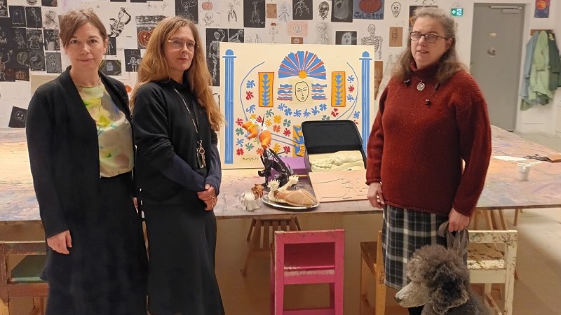 Tre vita medelålders kvinnor och en ledarhund, grå storpudel, står framför några taktila miniatyrmodeller av större konstverk. Exempelvis tavlan ”Apollon” av Matisse, detalj ur ”Paradiset” av Niki de Saint Phalle och ”Wilhelm Tellls gåta” av Dalí.