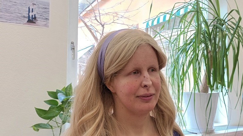 En ung kvinna med ljus hy och fräknar, och långt blont hårsvall som hålls bakåt av ett ljust lila hårband. Hon sitter vid ett fönster med gröna växter i.