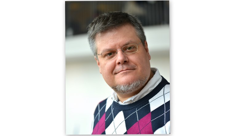 Man med grått hår och skägg på hakan och mösntrad tröja.