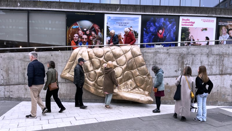 6 människor i alla åldrar och kön står framför ett konstverket föreställande ett täcke i guld. Täcker hänger ner över en ramp vid Sergels torg.