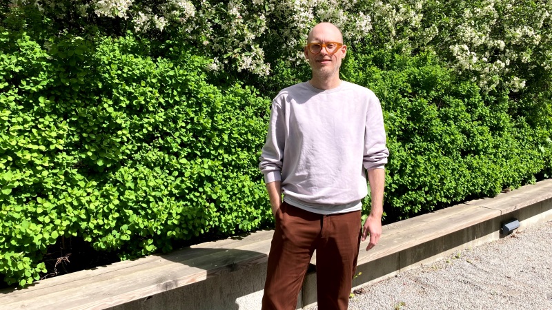 Vit, kal, medelålders man med orangebruna glasögon, grå tröja, bruna byxor. Står vid grönt buskage. Sommarsol.