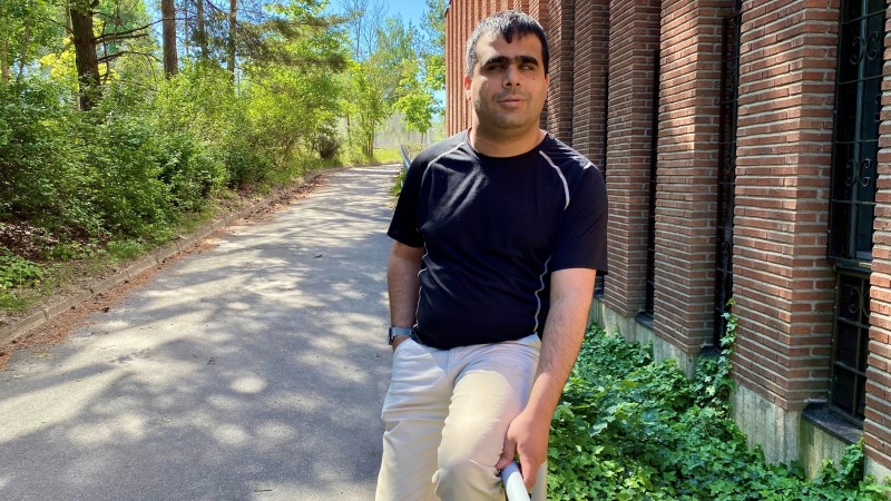 En yngre man med mörk hy och svart kort hår, svart t-shirt, beiga byxor. Han sitter på ledstång vid sidan av promenadväg. Lummig grönska på ena sidan, hög röd tegelbyggnad på den andra. Solig sommardag.