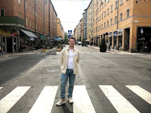 Yngre vit man med kort blont hår och mustache, beige jacka, vit t-shirt, blå jeans, vita sneakers. Står på Skånegatan.