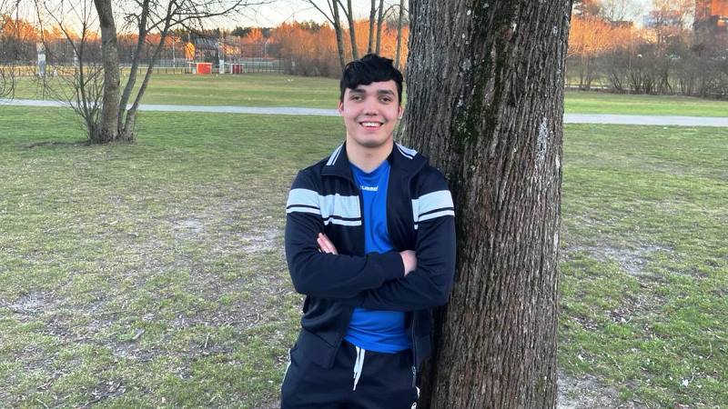 17 årig kille med mörkbrunt kort hår, står vid ett träd. Har svart träningsjacka, blå tröja och svarta träningsbyxor.