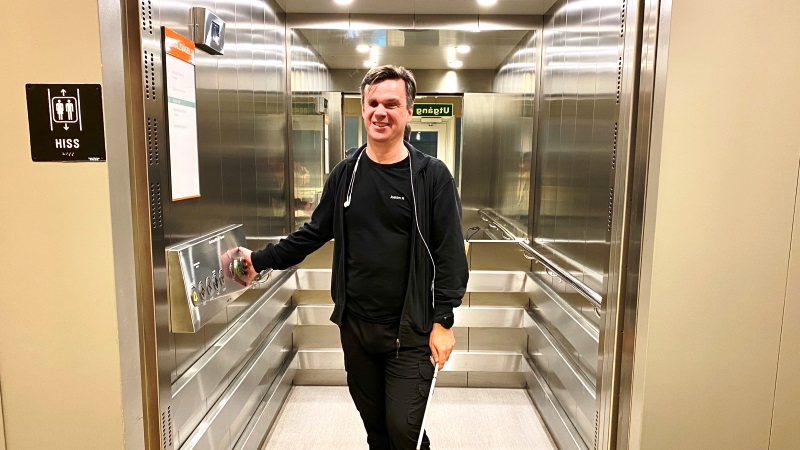 Medelålders ljushyad man med kort brunt hår, svarta kläder och vit käpp. Står i hiss med spegelväggar.