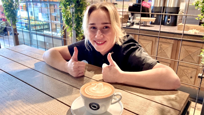 ljus, blond ung leende kvinna i svart blus, lutar sig över bord med en kopp cappucino, hon gör två tummar upp. Är på kafé.